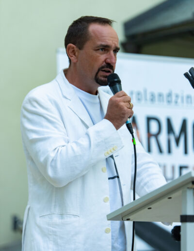 Roland Zingerle spricht bei einer Veranstaltung im Jahr 2019, gekleidet in einem weißen Sakko und hält ein Mikrofon in der Hand.