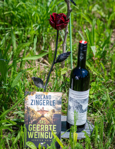 Ein Buch mit dem Titel "Das Geerbte Weingut" von Roland Zingerle, eine dekorative Rose und eine Flasche Hochosterwitzer Schlosswein im Gras.